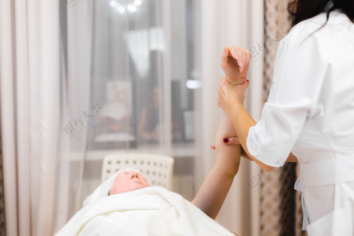 治疗一位年轻的女顾客脸上戴着海藻酸钠面膜 躺在美容台上 美容师做手工按摩女人面霜面部