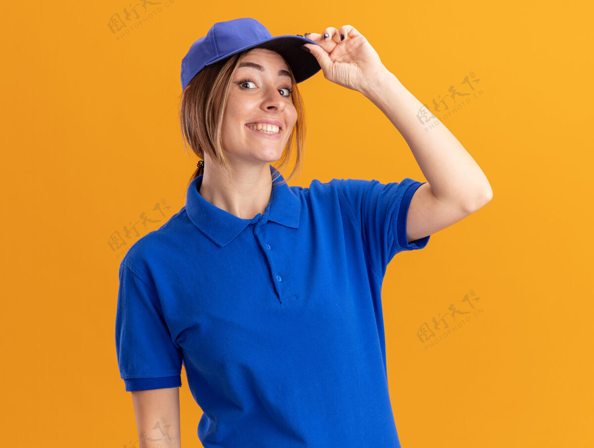 漂亮身着制服的年轻漂亮的女送货员微笑着把手放在橘色墙上的帽子上脸微笑站着