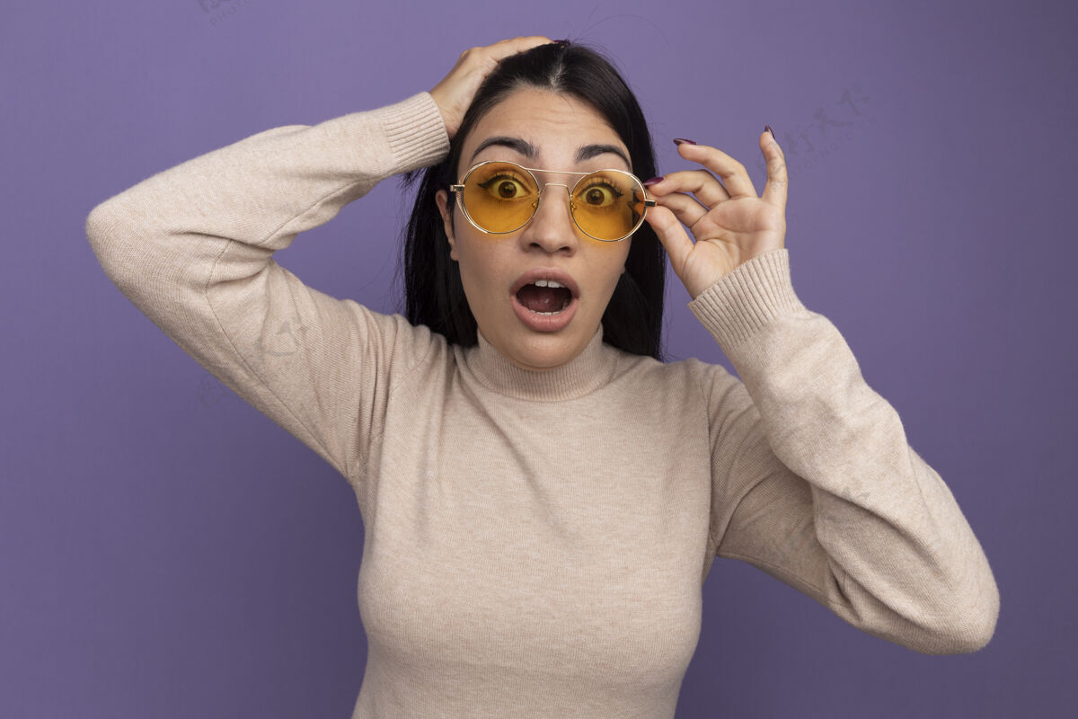 脸兴奋的黑发美女戴着太阳镜 把手放在头上 看着隔离在紫色墙上的前面感觉表情姿势