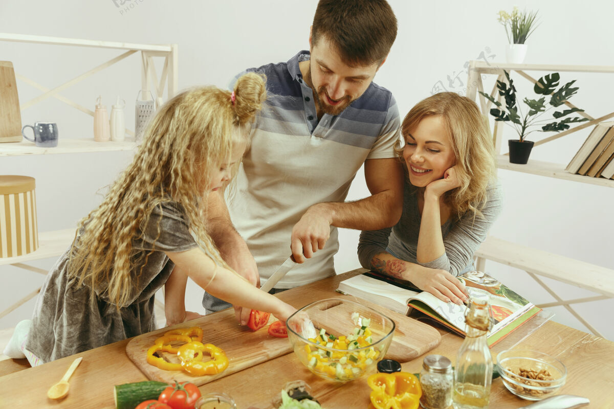 年轻可爱的小女孩和她美丽的父母正在家里的厨房里切菜 一边微笑着做沙拉家庭生活理念食物夫妇健康