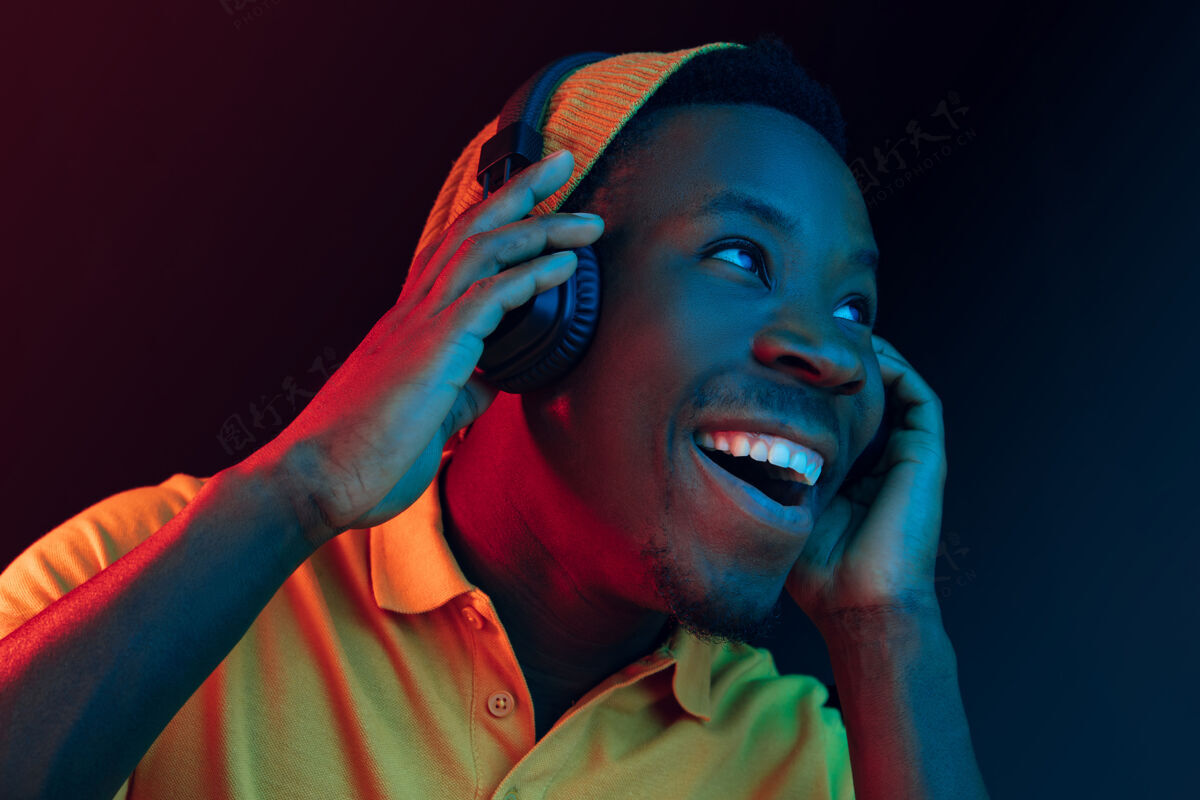 微笑年轻帅气快乐的时髦男人在霓虹灯下用耳机在黑工作室听音乐迪斯科舞厅 夜总会 嘻哈风格 积极的情绪 面部表情 舞蹈概念迪斯科球迷牙买加