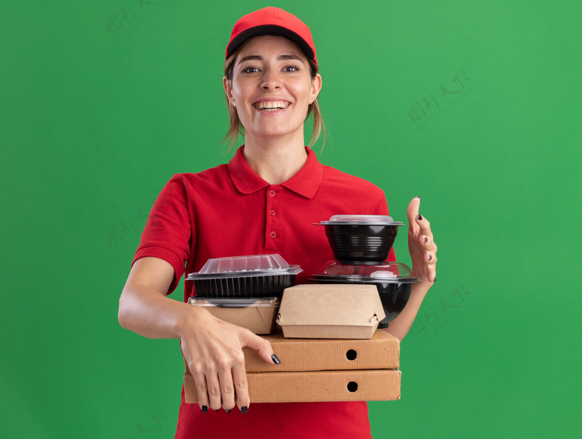 脸微笑着的年轻漂亮的女送货员穿着制服拿着纸食品包装和比萨饼盒上的容器隔离在绿色的墙壁上食物制服站着