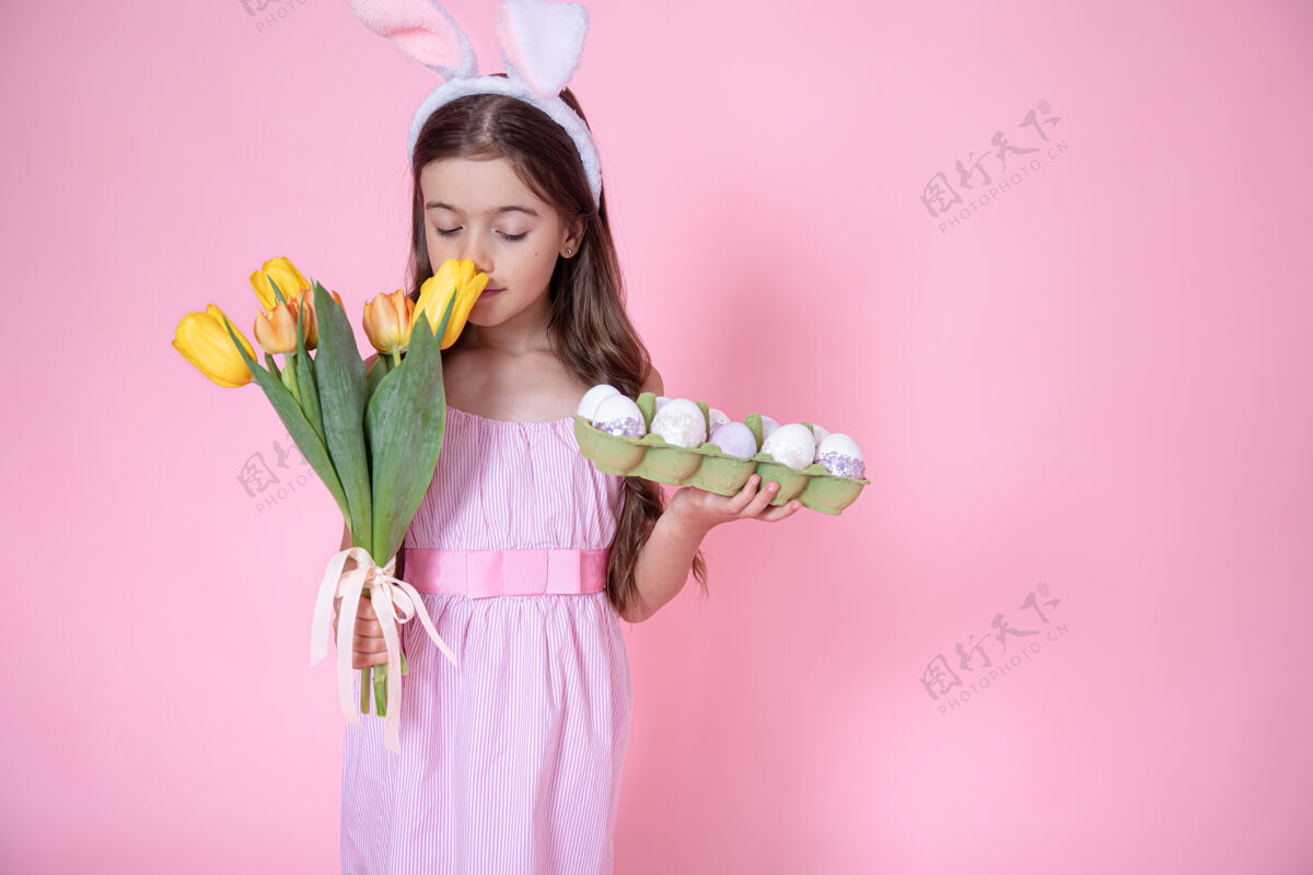 兔子一个有复活节兔子耳朵的小女孩 手里拿着一盘鸡蛋 在粉色工作室背景上嗅着一束郁金香托盘鸡蛋摄影棚