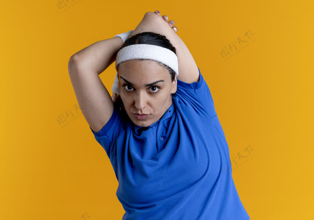 手臂年轻自信的白人运动女性戴着头带和腕带 将手臂放在背后 橙色背景上 留有复制空间头带佩戴运动