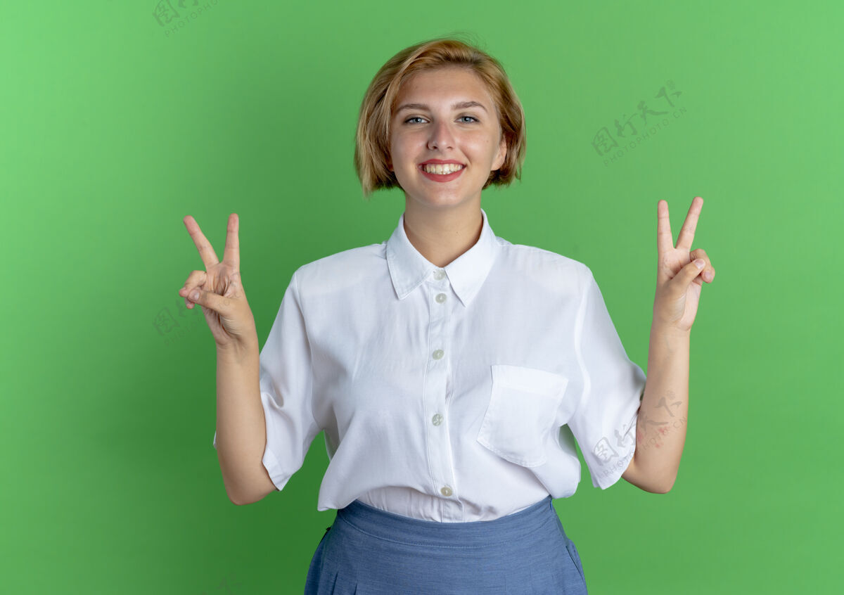 手年轻的微笑的金发俄罗斯女孩手势胜利手势与两只手在绿色背景与复制空间隔离金发年轻胜利