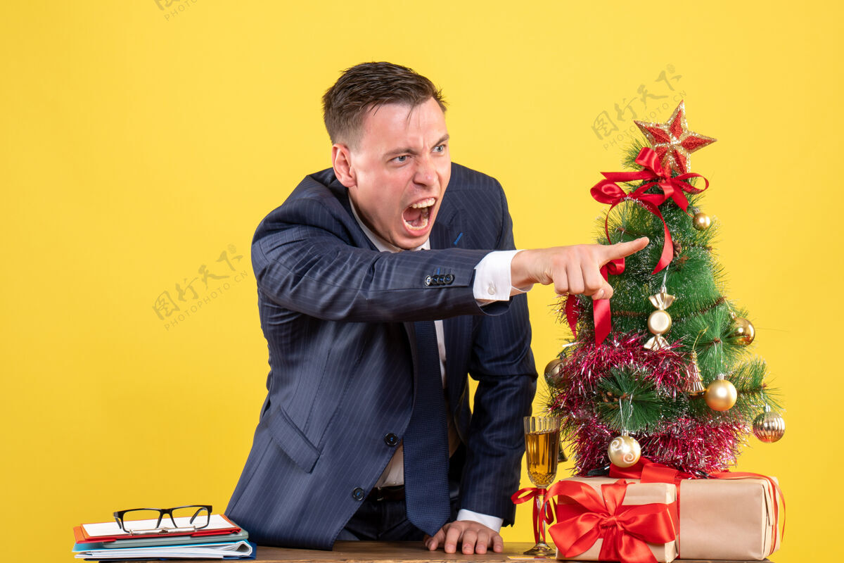 树前视图愤怒的男人对着站在圣诞树旁桌子后面的人大喊大叫 黄色背景的礼物生活前面男性