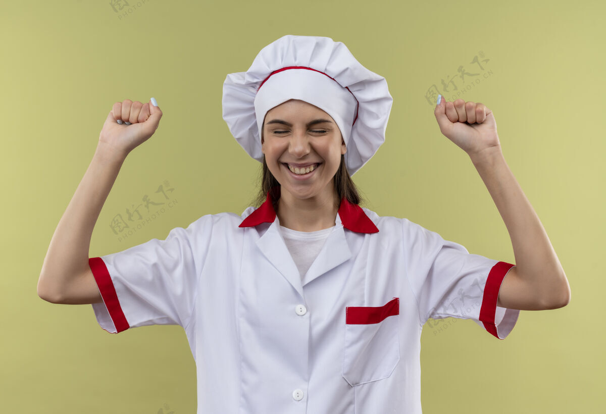 拳头身着厨师制服的年轻快乐的白人厨师女孩举起拳头孤立地站在绿色背景和复制空间上厨师制服烹饪