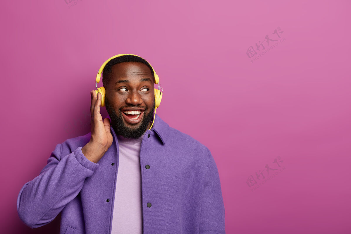 音频快乐的非洲裔美国人喜欢戴着耳机唱歌 听悦耳的旋律 侧目 心情愉快 穿着紫色夹克业余时间剃胡子男性