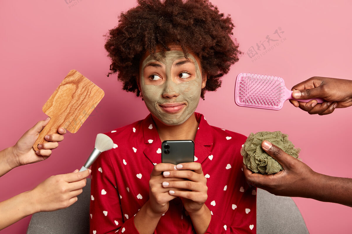 刷子体贴的非洲裔年轻女子一边若有所思地看着 一边在美容过程中用手机上网搜索 脸上涂上了粘土面膜睡衣打字海绵