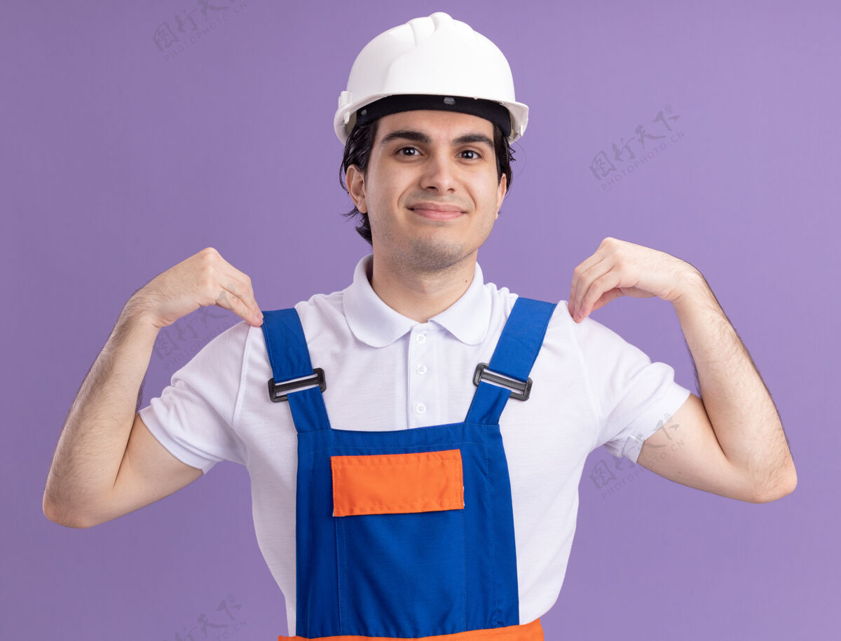 触摸身穿施工制服 头戴安全帽的年轻建筑工人站在紫色的墙上 面带微笑 举起双手抚摸着肩膀站立快乐脸