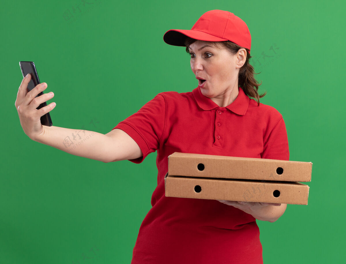 站着身穿红色制服 头戴鸭舌帽的中年送货妇女拿着披萨盒 站在绿色的墙上 看着智能手机 既惊讶又困惑女人拿着中年