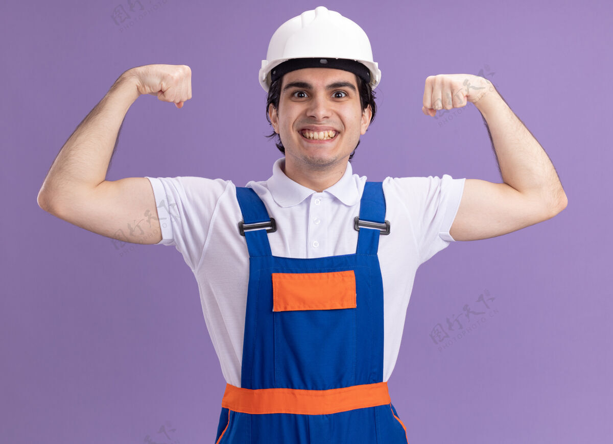 站立年轻的建筑工人身着建筑制服 戴着安全帽 笑容可掬地举起拳头 像一个胜利者站在紫色的墙上年轻微笑拳头