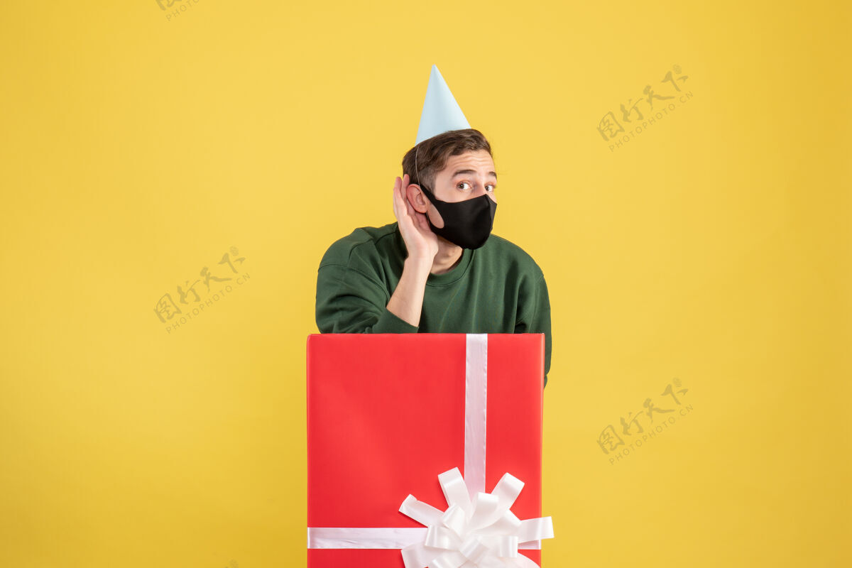 大正面图戴派对帽的年轻人站在黄色背景的大礼盒后面 听着什么东西礼物快乐人