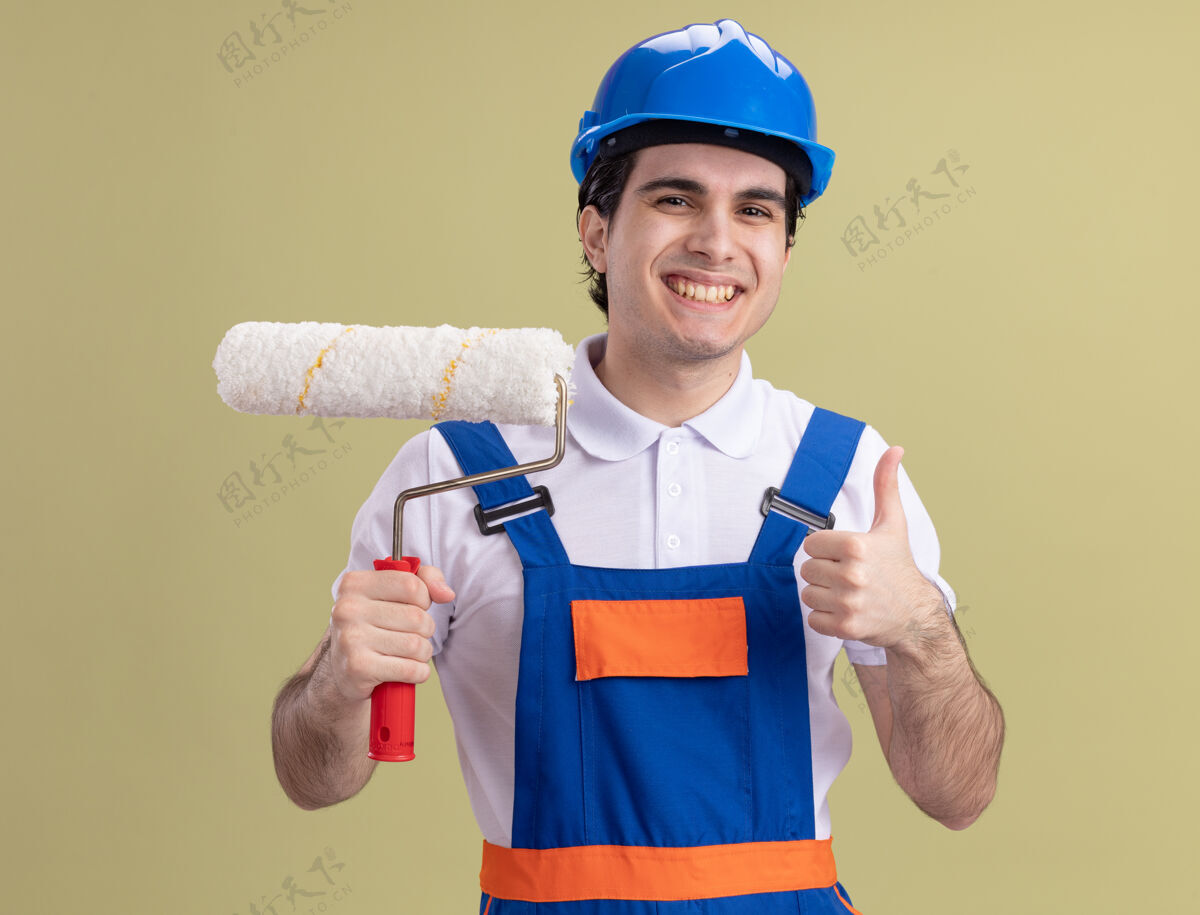 建设者年轻的建筑工人穿着建筑制服 戴着安全帽 拿着油漆辊 快乐而积极地看着前面 竖起大拇指站在绿色的墙上快乐拇指年轻