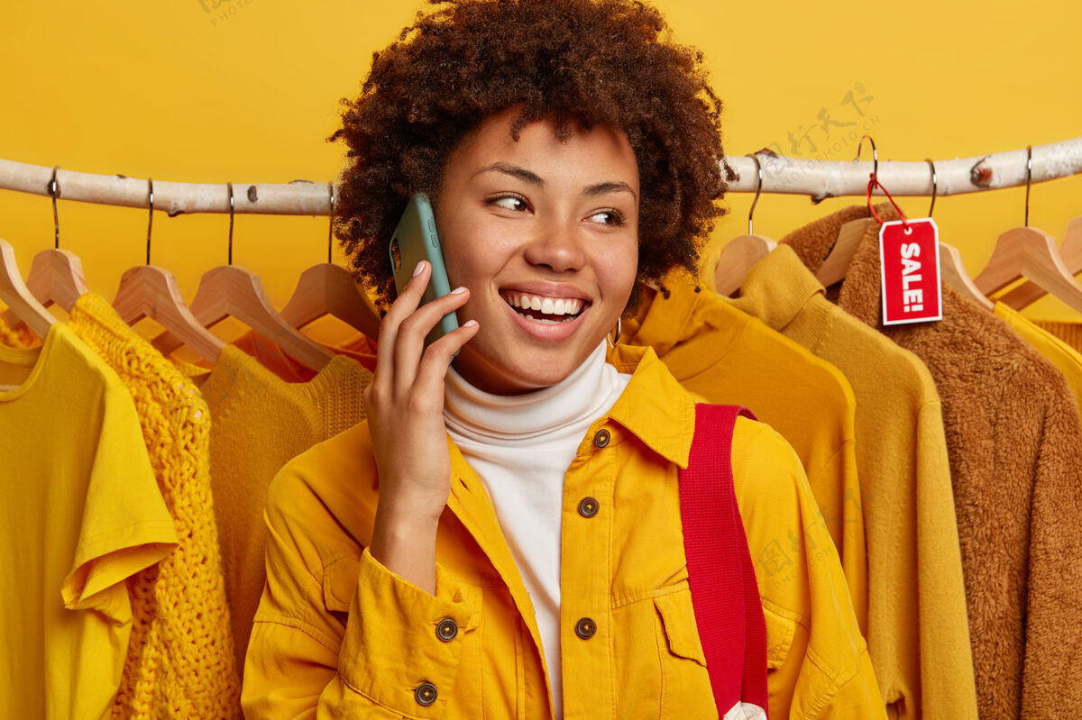 服装开朗的非洲裔美国妇女在手机上交谈 在商店里检查一些衣服 在衣架上摆姿势 分享商店里的销售情况电话美式购物狂