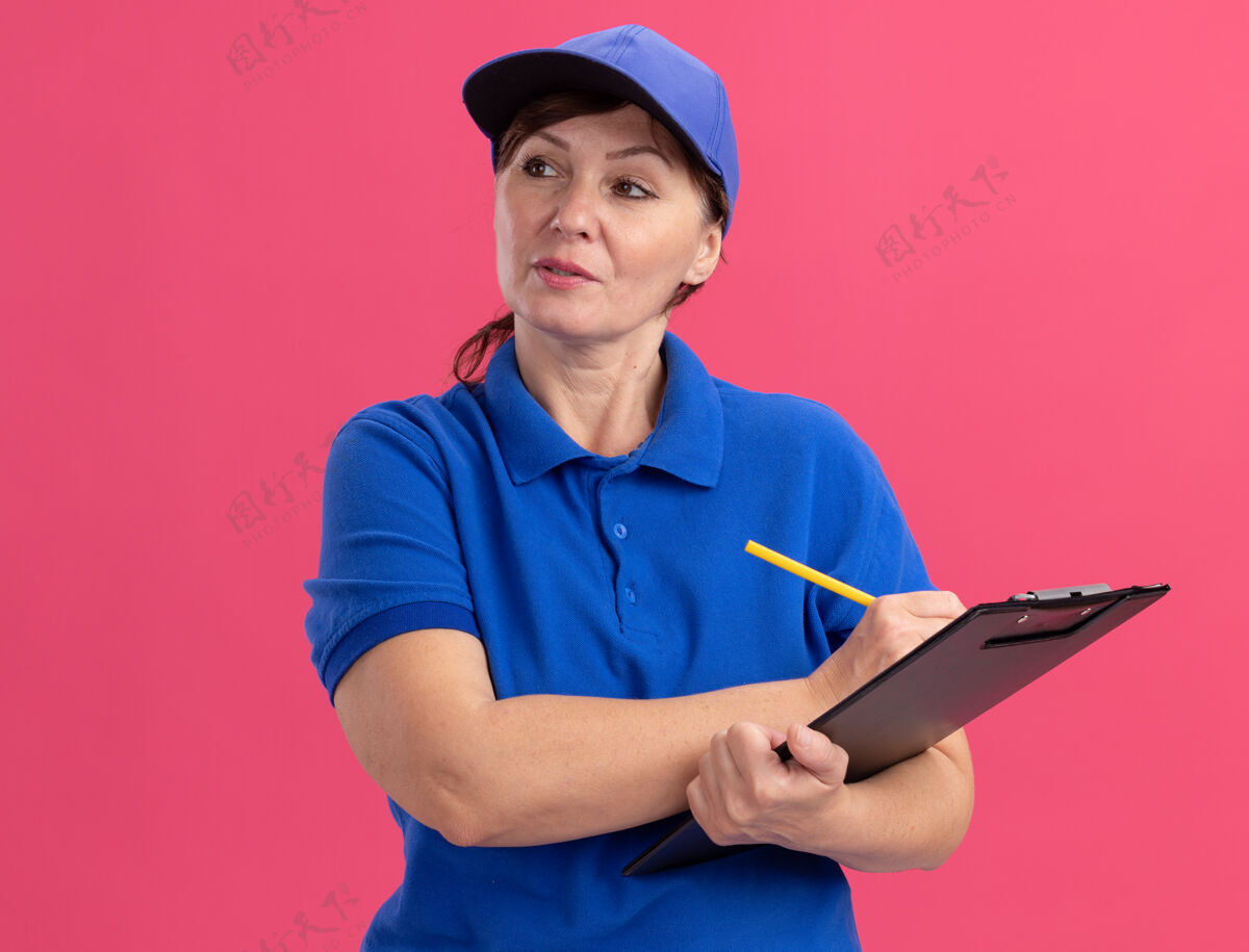 女人身穿蓝色制服 头戴鸭舌帽的中年女送货员站在粉红色的墙上 手里拿着剪贴板和铅笔 神情严肃自信地看着一旁快递严肃表情