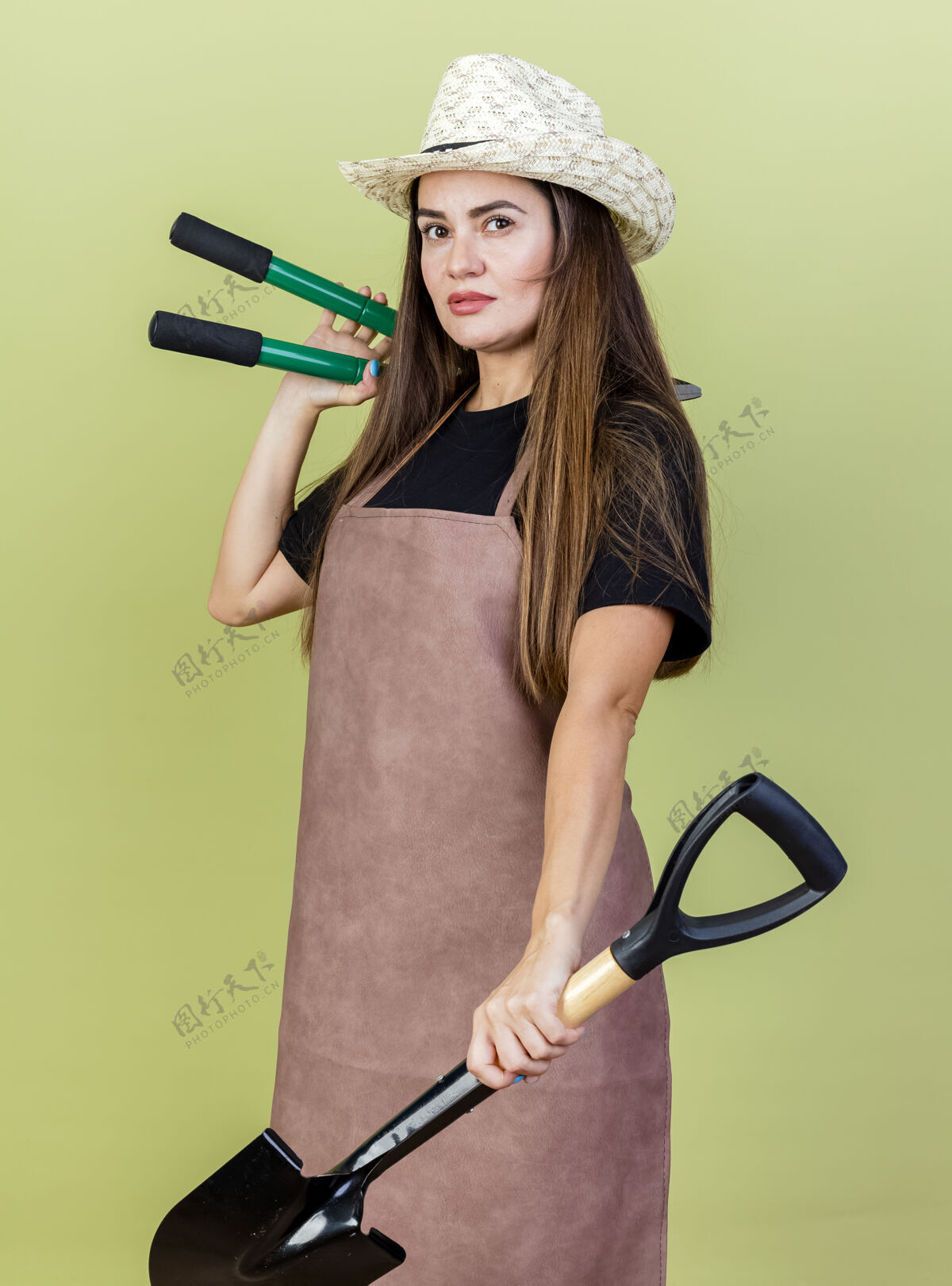举行自信美丽的园丁女孩穿着制服 戴着园艺帽 手持铁锹 肩上放着剪子 橄榄绿的背景上与世隔绝女孩园艺黑桃