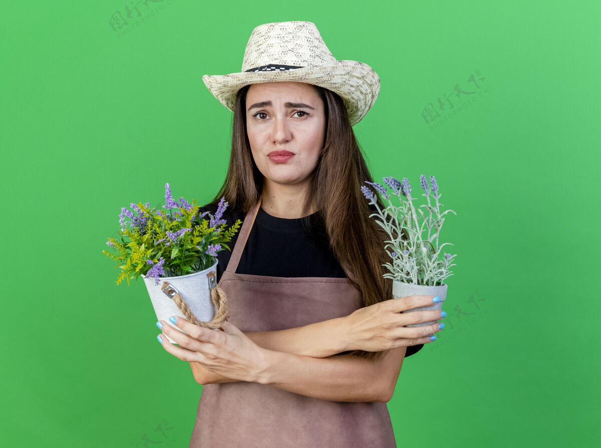关心关心美丽的园丁女孩穿着制服 戴着园艺帽 在绿色的背景下 在花盆里拿着和交叉的花交叉穿花园
