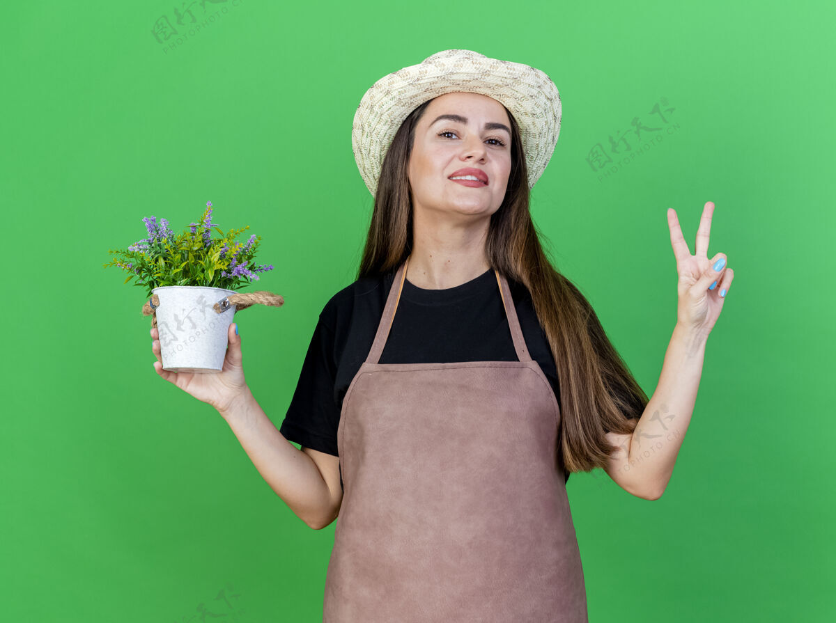 女孩微笑美丽的园丁女孩穿着制服 戴着园艺帽 在花盆里拿着花 在绿色的背景上显示出与世隔绝的和平姿态花园微笑穿