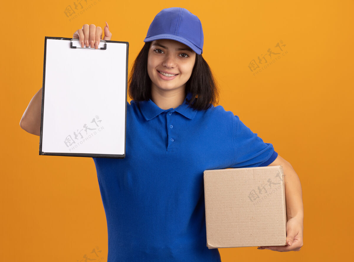 举行身穿蓝色制服 戴着帽子的年轻送货女孩站在橙色的墙上 手里拿着一个纸板箱 上面有空白页的剪贴板 她微笑着自信地站着女孩帽子交货