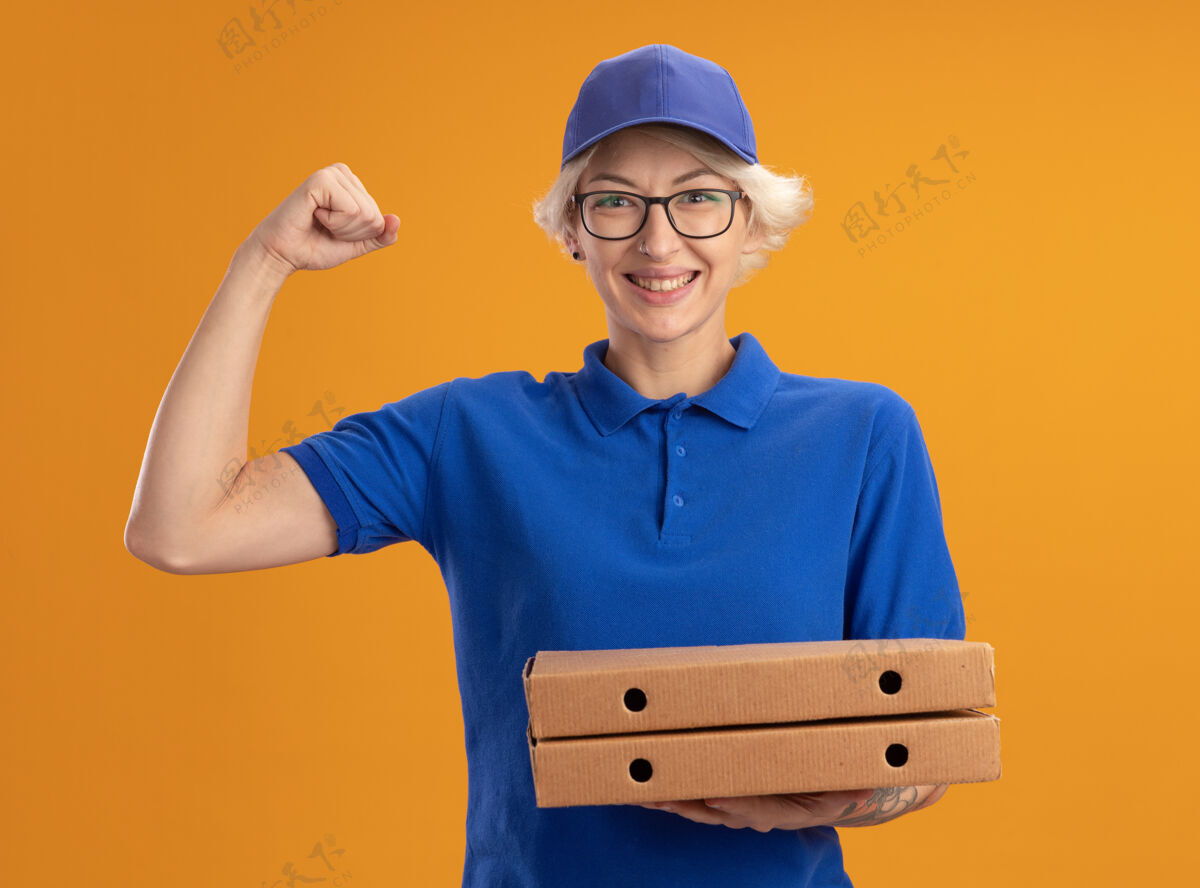 制服身着蓝色制服 戴着眼镜 拿着比萨饼盒的年轻送货员快乐而自信地握紧拳头越过橙色的墙壁戴着披萨盒子