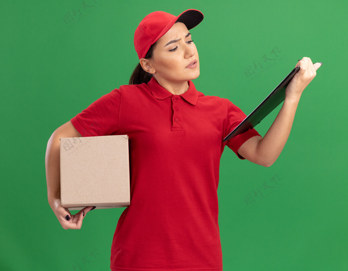 女人穿着红色制服 戴着帽子 拿着剪贴板的年轻女送货员拿着纸板箱 严肃地看着剪贴板站在绿色的墙上严重纸板剪贴板