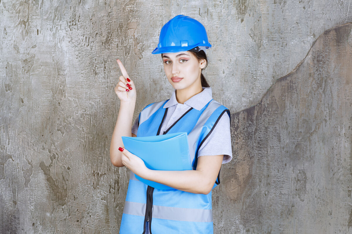 演示身穿蓝色制服 头戴安全帽的女工程师手持蓝色报告夹 指着周围的人项目施工姿势