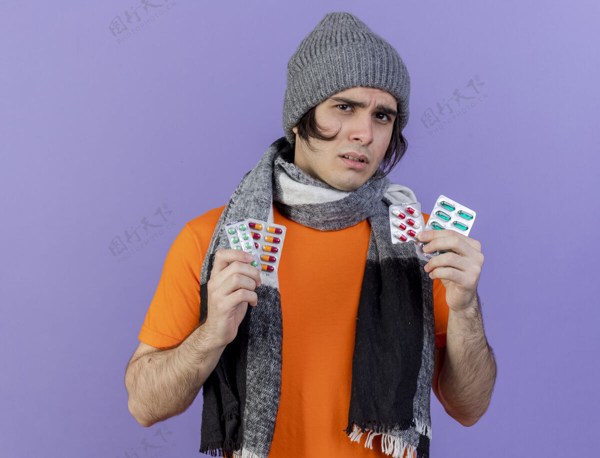生病困惑的年轻病人戴着冬天的帽子 戴着围巾 拿着紫色背景上孤立的药丸帽子围巾迷糊