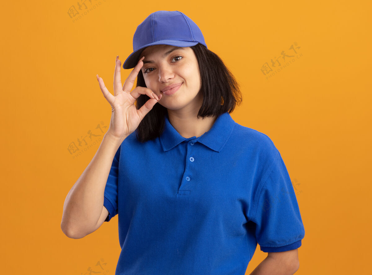 帽子身穿蓝色制服 头戴鸭舌帽的年轻送货女孩 做出沉默的手势 就像站在橘色墙上的拉链合上嘴巴拉链站立关闭