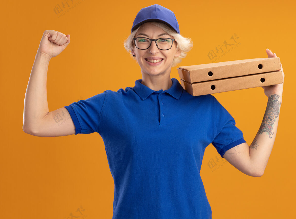 提高身着蓝色制服 戴着眼镜 手持披萨盒的年轻送货员在橙色的墙上高举着快乐的拳头拿着欢呼眼镜