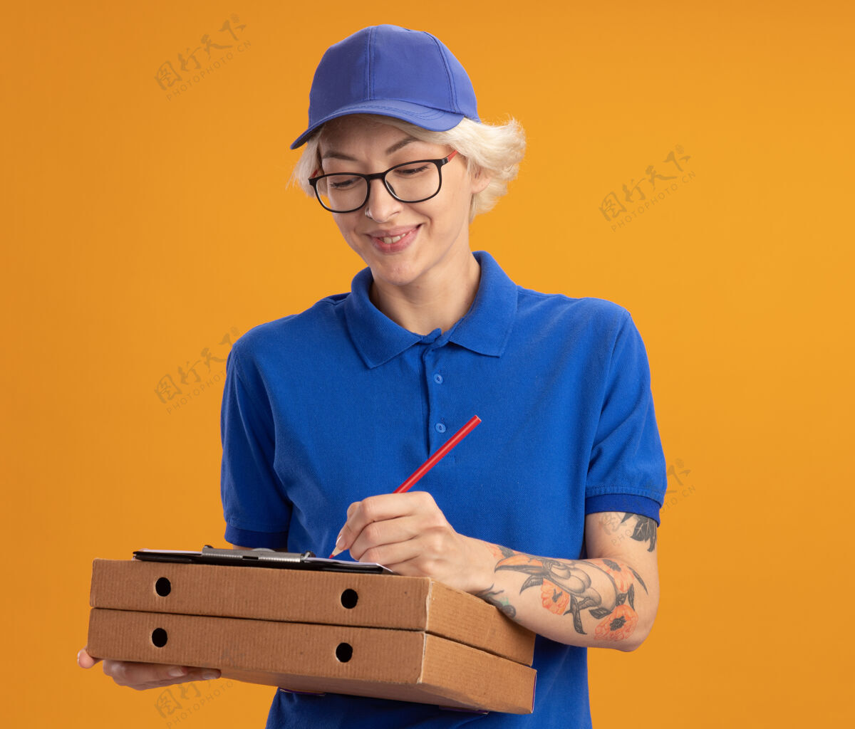女人身穿蓝色制服 戴着眼镜 拿着披萨盒和空白页的剪贴板 用铅笔在橙色墙上微笑着写着什么的年轻女送货员空白眼镜穿