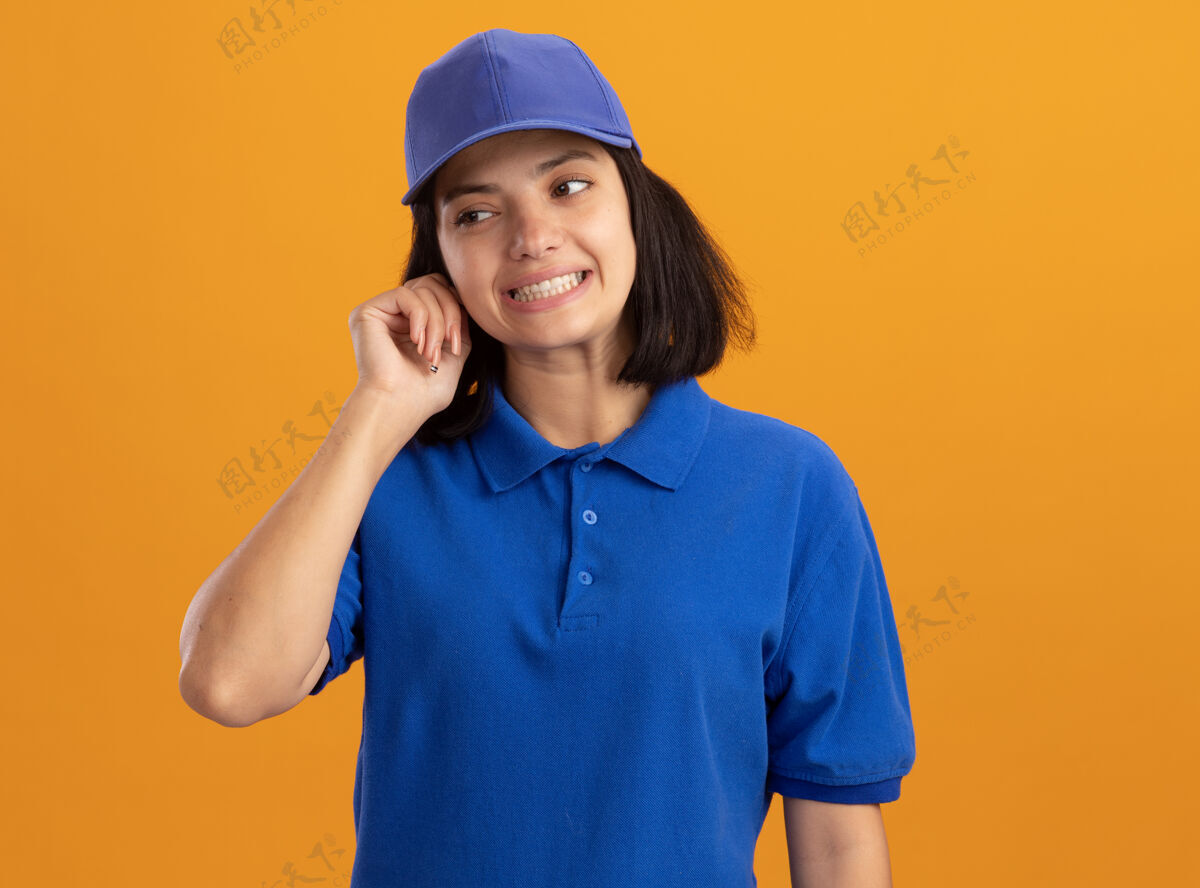 帽子身穿蓝色制服 戴着帽子的年轻送货员站在橙色的墙上 困惑地向一旁望去制服站在一边交货