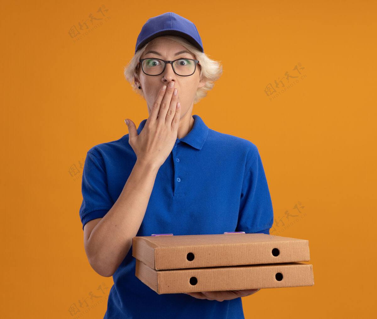 眼镜身穿蓝色制服 戴着眼镜 手持比萨饼盒的年轻送货员被震惊地用手捂住嘴巴 越过橘色的墙壁披萨制服送货