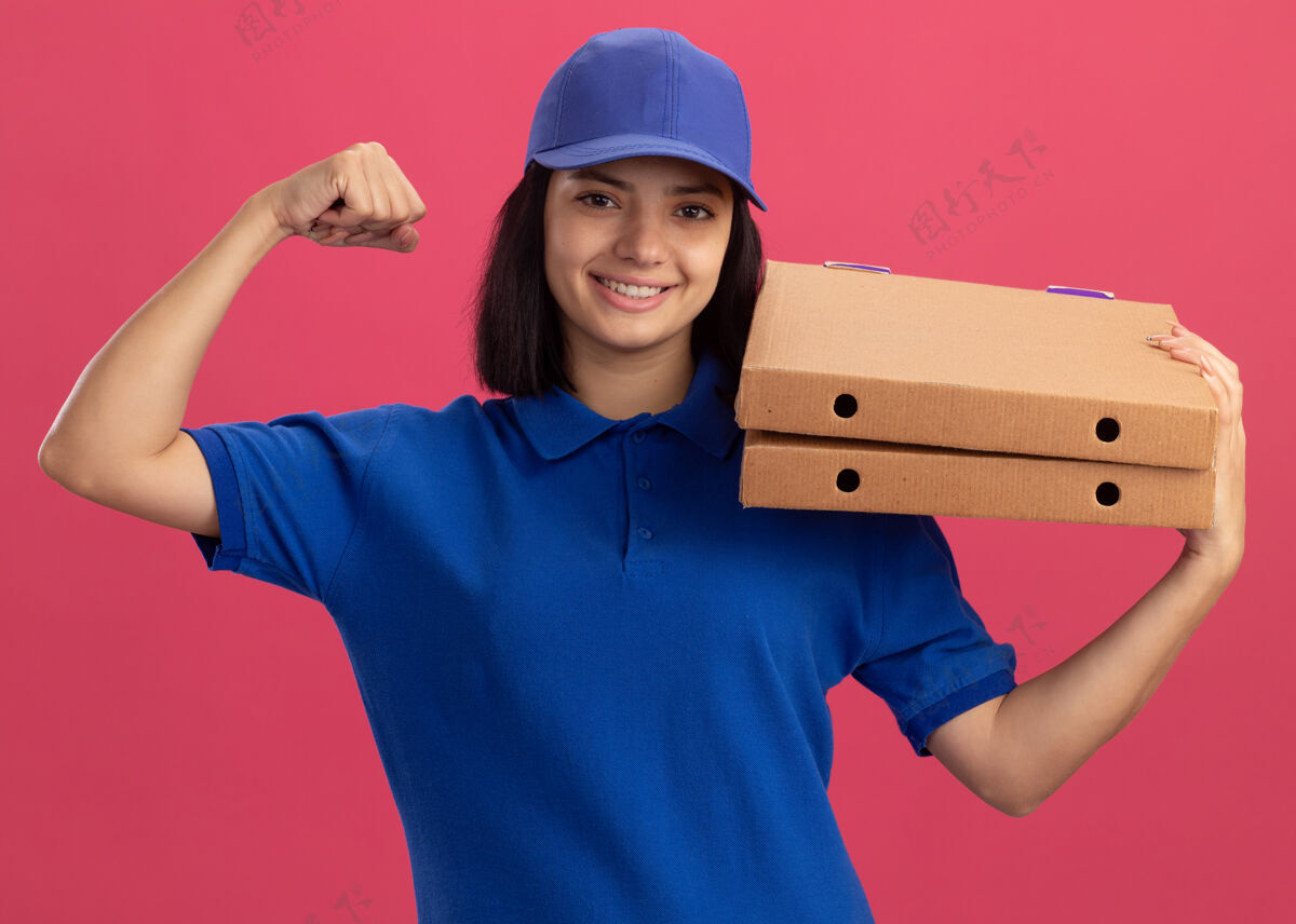 送货身穿蓝色制服 头戴鸭舌帽 手持披萨盒的年轻送货员举起拳头 站在粉红色的墙上 像一个胜利者一样高兴地微笑着举行披萨微笑