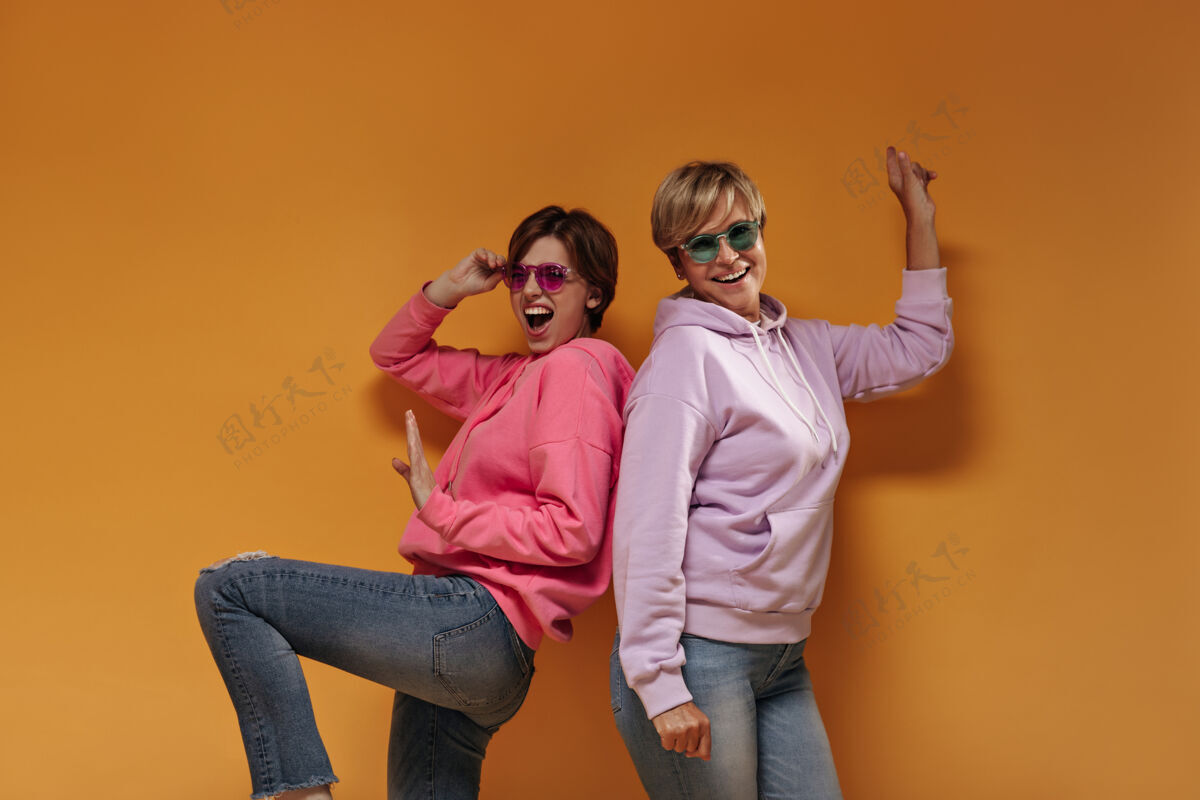 太阳镜带着墨镜 穿着粉色运动衫的情绪化女孩笑着和身着淡紫色连帽衫 橙色背景的老太太合影女连帽衫老年人