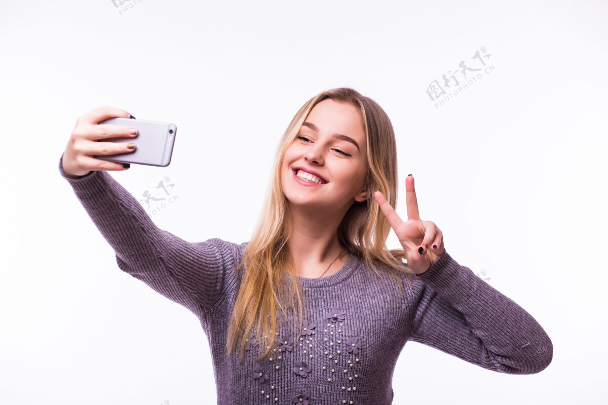 微笑一个微笑的可爱的女人在智能手机上自拍的照片被隔离在白色的墙上胜利成人聪明