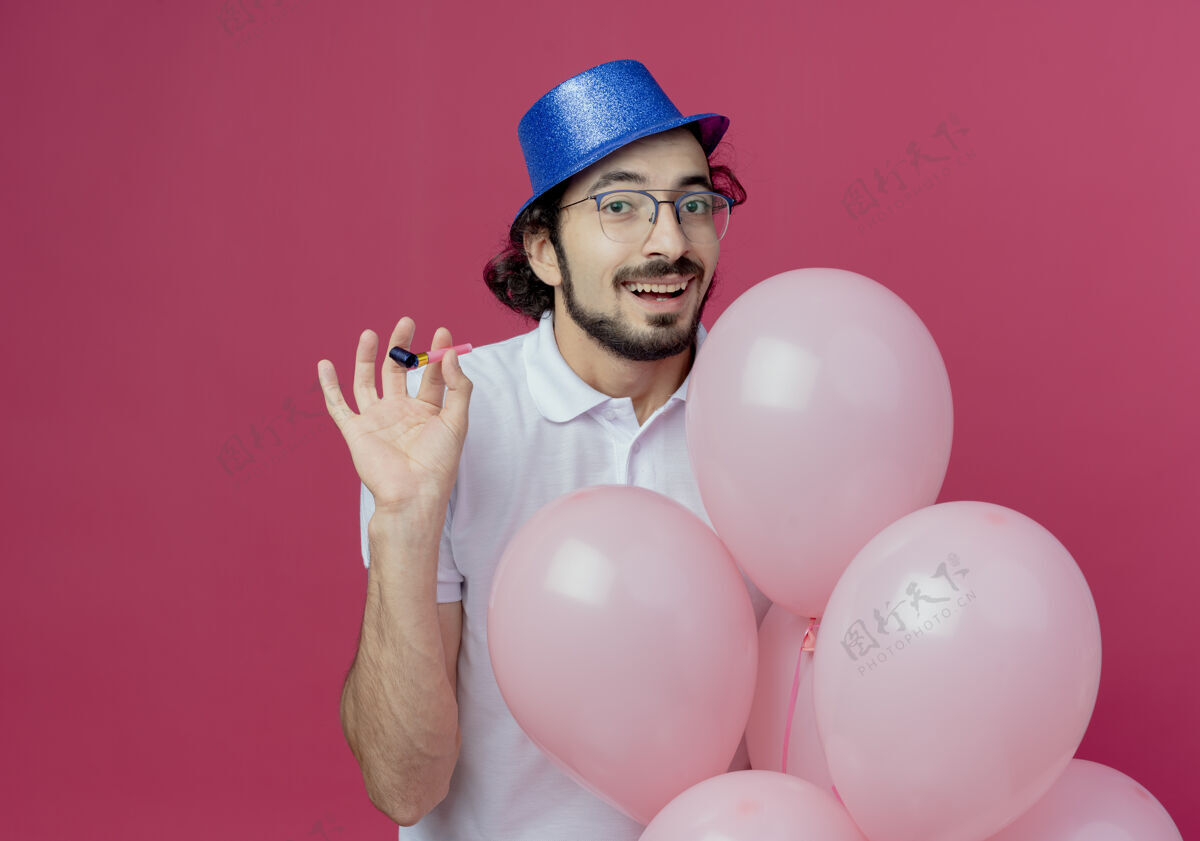 帅哥高兴的帅哥戴着眼镜 戴着蓝色的帽子 手持气球 吹着隔离在粉色背景上的口哨手持请戴上口哨