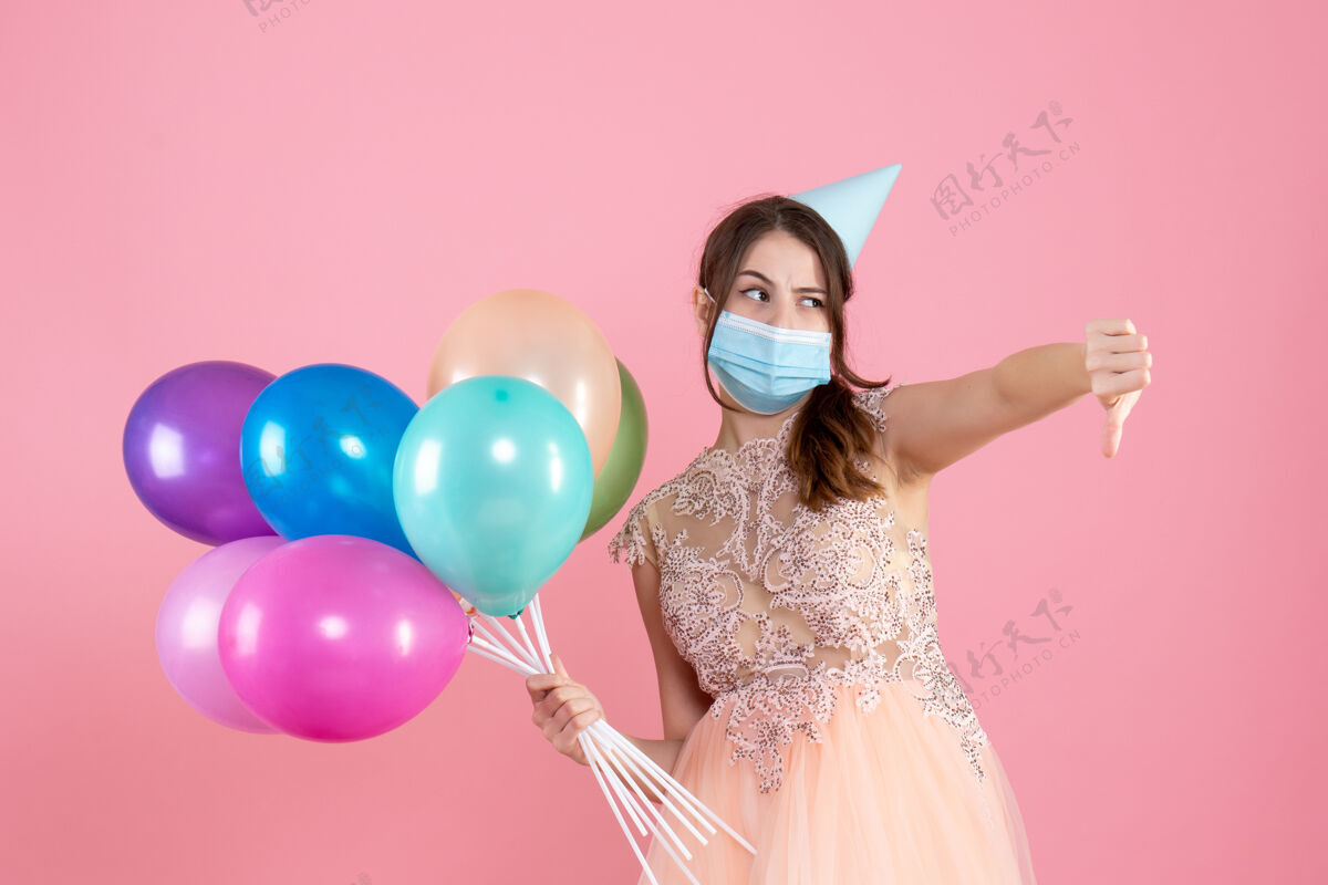 粉红色前视图困惑的女孩 戴着派对帽和医用面罩 拿着五颜六色的气球做着拇指朝下的手势标志乐趣困惑的女孩