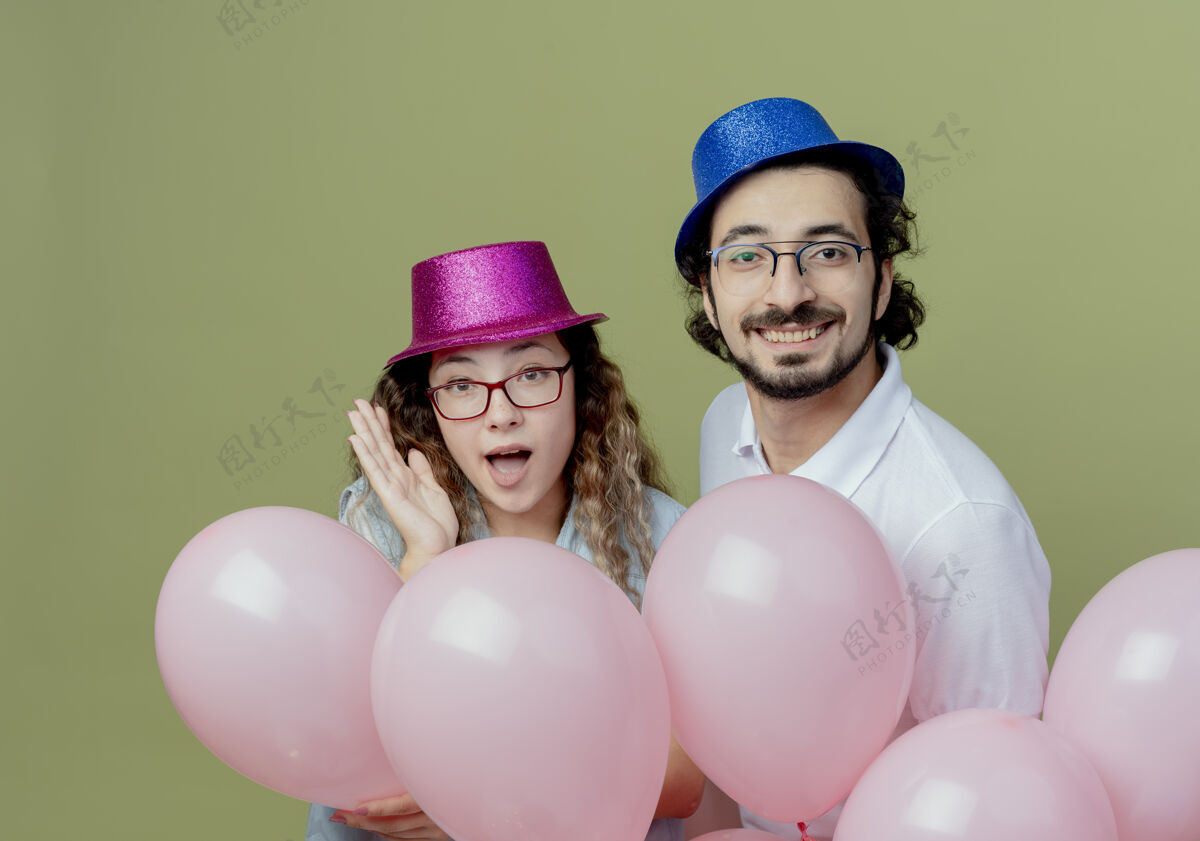 气球快乐的年轻夫妇戴着粉红色和蓝色的帽子站在橄榄绿背景上孤立的气球后面穿上情侣快乐