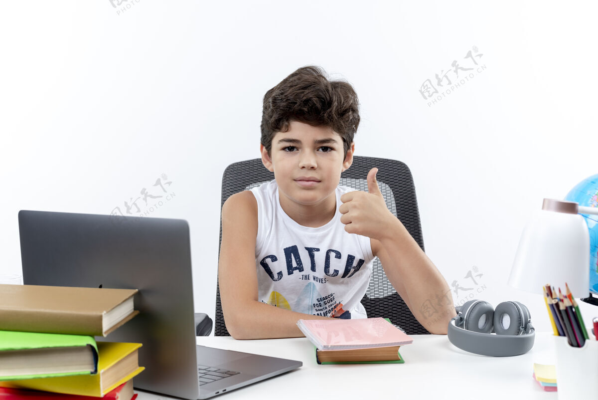 学校高兴的小男孩坐在书桌旁 手里拿着学习工具 他的大拇指孤立地竖立在白色的背景上求求你书桌拇指