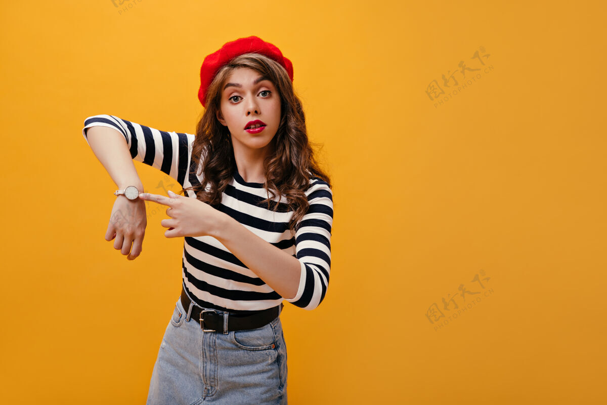牛仔裙戴贝雷帽的卷发女孩指着手表穿着条纹毛衣的卷发现代年轻女子在孤立的背景下摆姿势复制帽子紫色