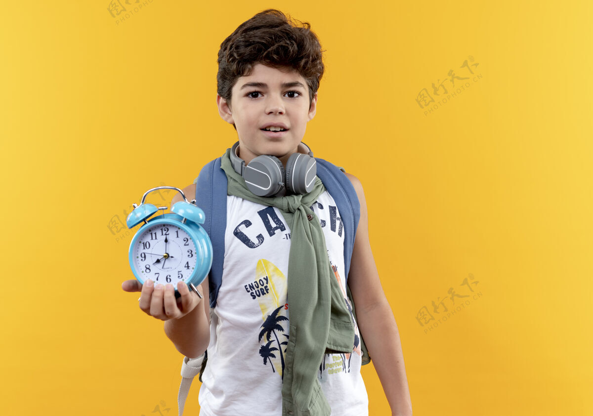 印象印象深刻的小男孩穿着背包和耳机举行黄色背景上的闹钟隔离包抱背