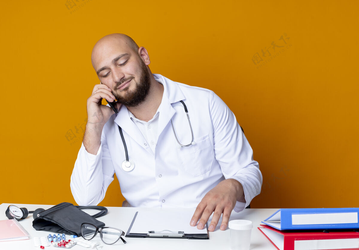 医生高兴的年轻秃顶男医生穿着医用长袍和听诊器坐在办公桌前 手持医疗工具 在橙色背景下讲电话电话长袍穿