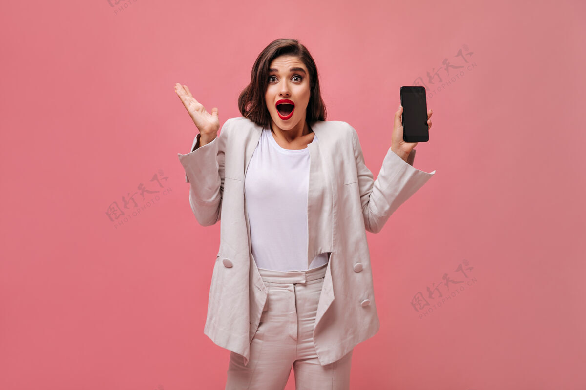 优雅一位穿着棉服的震惊女士在粉色背景上演示手机一个穿着白衣服 嘴唇明亮的惊讶女人手里拿着电话女孩成人肖像
