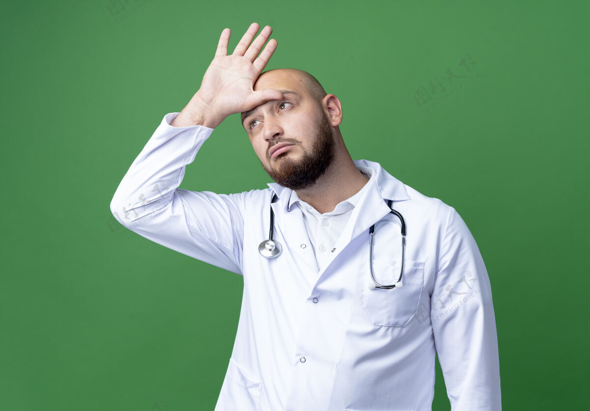 思维看着一边想着的年轻男医生 穿着医用长袍 听诊器 手放在头上 隔离在绿色的背景上听诊器长袍手