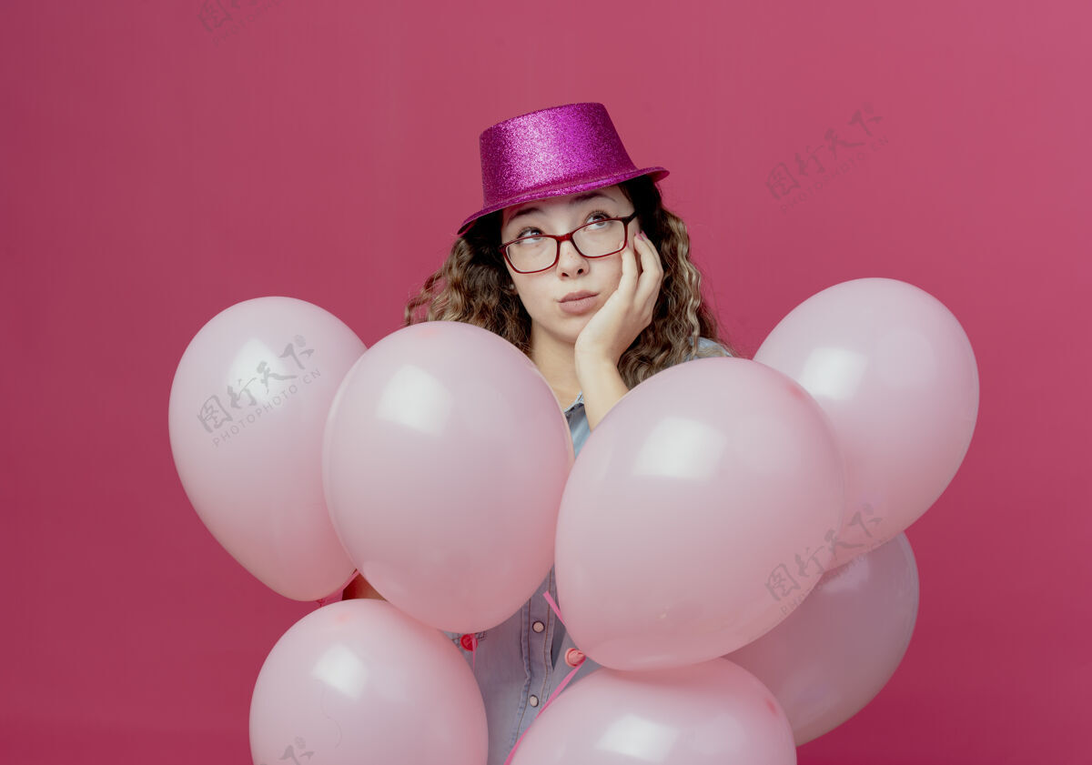 脸颊看着侧面思考的戴着眼镜和粉色帽子的年轻女孩站在气球中间 手放在脸颊上 孤立在粉色背景上思考女孩穿