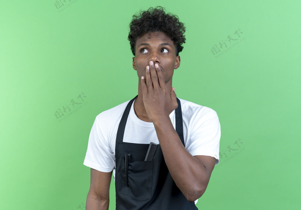 穿看着旁边惊讶的年轻美国黑人男理发师穿着制服捂着嘴 手孤立在绿色背景上男性理发师封面
