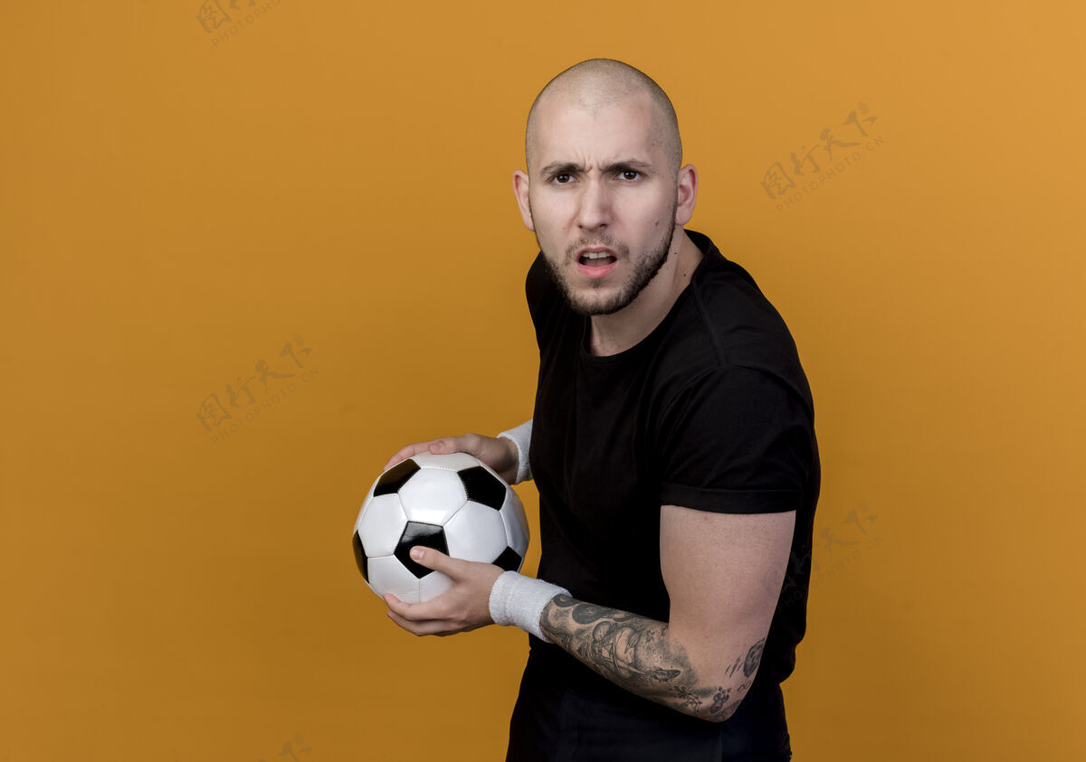 运动困惑的年轻运动男子戴着手环拿着球孤立在橙色背景与复制空间持有腕带年轻