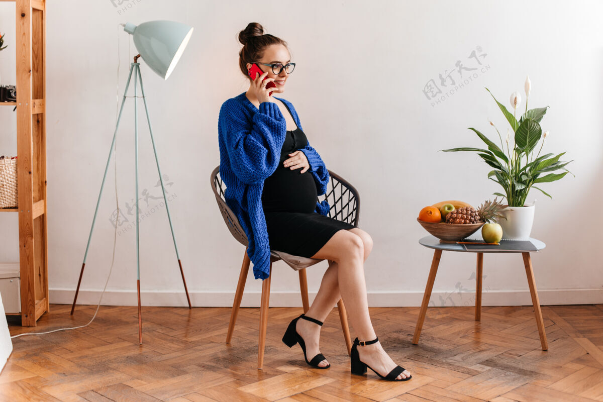 室内身着黑色连衣裙和蓝色开衫的迷人女士坐在木椅上讲电话戴眼镜的孕妇摸肚子笔记本坐着房间