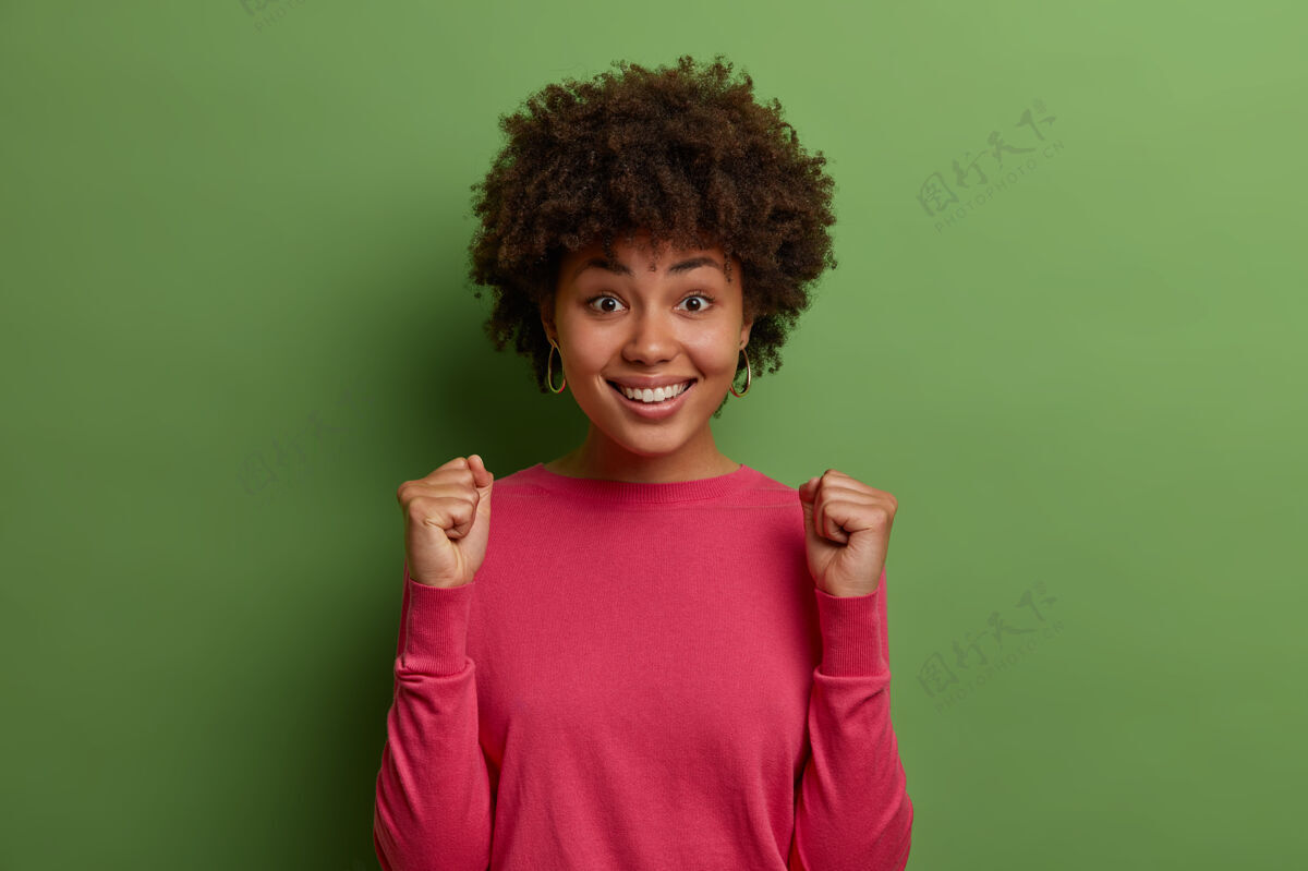 冠军喜气洋洋的卷发女人等待重要的结果 握紧拳头庆祝 津津乐道的好消息 高兴地微笑 高兴地实现目标 穿着粉红色的套头衫 孤立在绿色的墙上笑肖像卷发
